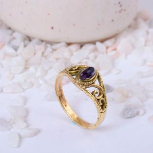 May include: A gold-coloured ring featuring a dark purple oval gemstone. The ring has an ornate, filigree design and a textured band. The gemstone is set in a bezel setting. The ring is on a white surface.