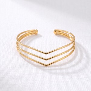 May include: A gold-toned cuff bracelet featuring three parallel bands that converge into a central V-shaped point. The open-ended design includes small rounded caps at each terminal end, creating a sleek, layered appearance with a polished finish.