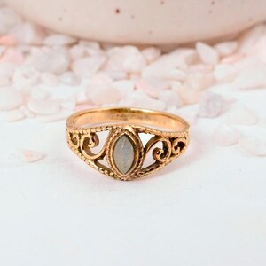 May include: A gold-colored ring with an oval, light-colored stone at the center. The ring features an ornate, filigree design around the stone and band. The ring is set against a white background with small, light-colored pebbles.