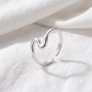 May include: A polished silver-tone ring featuring an open, wavy, or fluid design. The band has a smooth, reflective surface with a continuous, undulating curve that creates an asymmetrical shape, forming a gap at the top of the ring structure.