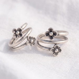 May include: Two silver-toned rings featuring a bypass spiral design. Each ring is adorned with two small, four-petaled flower motifs at the ends of the band. The metal has a matte, slightly oxidized finish, highlighting the floral details.