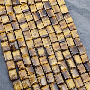May include: A collection of rectangular tiger's eye gemstone beads arranged in multiple vertical strands. Each bead features a polished, smooth surface with characteristic golden-brown chatoyant bands and dark brown inclusions throughout the stones.