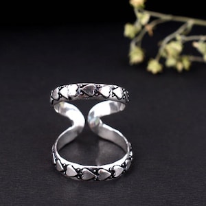 Puede incluir: Anillo doble para dedos de color plateado con diseño de corazones. El anillo presenta dos bandas conectadas por una sección curva. Las bandas están adornadas con pequeñas formas de corazón. El anillo se muestra sobre un fondo oscuro.