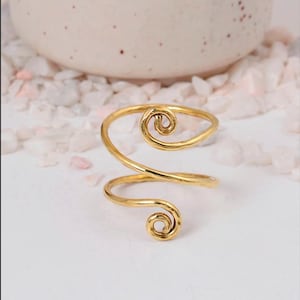 May include: A gold-colored ring with a spiral design. The ring features two swirling, scroll-like accents at the top and bottom, connected by a curved band. The ring is set against a white surface with small, light pink and white pebbles.