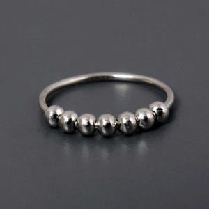 May include: A silver ring featuring seven small, spherical beads encircling the band. The ring's design is minimalist, set against a grey backdrop. The beads are evenly spaced around the ring.