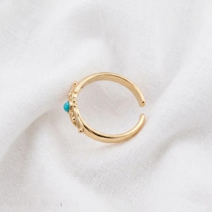 Può includere: Un anello aperto color oro con una piccola pietra turchese rotonda incastonata in una montatura in metallo decorativa e testurizzata. La fascia è liscia e circolare, progettata con un'apertura regolabile per diverse misure di dita.