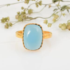 May include: A gold-colored ring featuring a large, light blue, rectangular gemstone. The gemstone is set in a gold bezel with a textured edge. The ring's band is also gold-colored, creating a cohesive and elegant design.