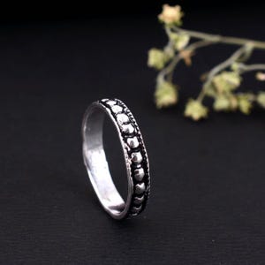 Puede incluir: Un anillo plateado con un diseño de cuentas alrededor de la banda. El anillo tiene un estilo sencillo y elegante, adecuado para el uso diario. El anillo se muestra sobre un fondo oscuro, resaltando su brillo metálico.