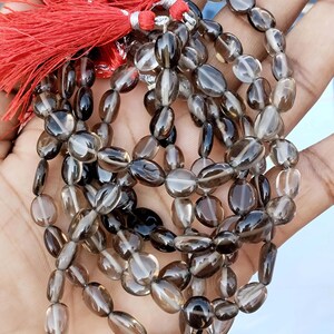 Può includere: Una collana di perle di quarzo affumicato con un fiocco rosso. Le perle sono di forma ovale e hanno un colore marrone affumicato traslucido.