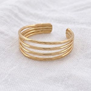 Op de afbeelding: Een goudkleurige ring met een ontwerp van meerdere banden, bestaande uit vier dunne, parallelle en gehamerde metalen draden. De ring heeft een open constructie voor aanpassing en toont een getextureerd, reflecterend oppervlak.