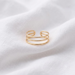 Könnte beinhalten: Ein goldfarbener Ring mit einem dreireihigen Design und einer glatten, polierten Oberfläche. Der Ring hat eine offene Struktur und zeigt drei parallele, dünne, kreisförmige Bänder, die an den Seiten verbunden sind und einen Lagen-Look erzeugen.