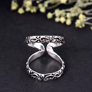 May include: A silver-colored ring with an open design. The ring features an intricate floral pattern around the bands. The two bands are connected by curved supports, creating a unique and stylish look. The ring is set against a dark background.