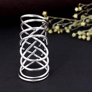 May include: A silver-colored finger ring with a unique design. The ring features a series of interconnected, curved bands that create an open, woven pattern. The ring is set against a dark background, with a hint of greenery in the upper right corner.