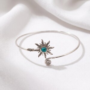 May include: A sterling silver cuff bracelet featuring a sun-shaped centerpiece with a round turquoise stone at its center. The metal band has a polished finish, a spiral end, and is clearly stamped with the number 925 to indicate silver purity.