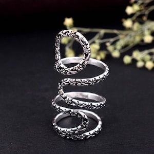 May include: A silver-colored, adjustable ring with a floral pattern. The ring wraps around the finger in a spiral design, with a loop at the top. The ring is made of metal and has an antique finish. The ring is a piece of jewelry.