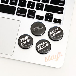 May include: Four black and white round buttons with the names of the different units of the South Korean boy band Stray Kids. The buttons read "Stray Kids", "3racha", "Vocalracha", and "Dance Racha". The buttons are on a white surface with the words "stay" and two pink hearts written in cursive.