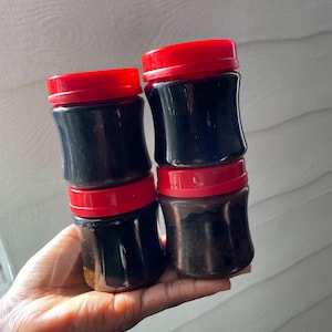 May include: Four small, clear jars with dark contents and bright red lids are stacked in a hand. The jars have a curved shape and are filled with a dark, viscous substance. The lids are a vibrant red, contrasting with the dark contents.