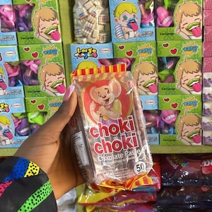 May include: A hand holds a packet of "choki choki" chocolate paste, with a cartoon mouse on the front. The background shows boxes of sweets with cartoon characters and the words "Large Tongue" and "Oops!" printed on them.