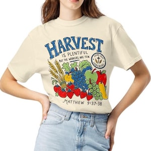 May include: A cream-colored t-shirt featuring a colorful graphic of wheat, grapes, strawberries, apples, and pears. The text reads: HARVEST IS PLENTIFUL BUT THE WORKERS ARE FEW. SEND OUT LABORERS. MATTHEW 9:37-38. The design uses blue, red, green, and yellow.