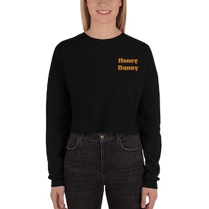 May include: Black cropped sweatshirt with long sleeves. The text "Honey Bunny" is embroidered in a gold color on the upper left chest area. The sweatshirt is made of a soft material.