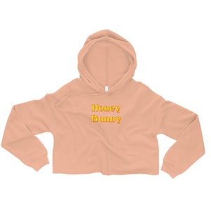 May include: A peach-colored cropped hoodie with a drawstring hood. The hoodie has the words "Honey Bunny" printed on the front in yellow and orange.