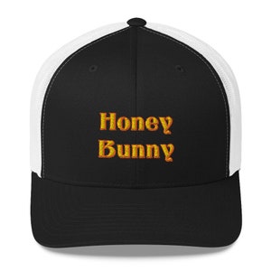May include: Black and white trucker hat with a black mesh back. The front of the hat has the words "Honey Bunny" embroidered in orange.