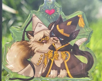 May include: A clear acrylic keyring featuring two cartoon cats dressed as wizards. One cat is light brown and the other is black, both with gold detailing. A red heart is above them. The keyring has a gold-coloured clasp.