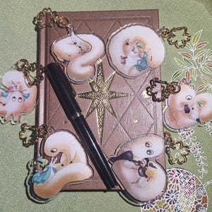 Può includere: Una collezione di portachiavi in acrilico con illustrazioni fantasiose di creature in varie pose. I portachiavi sono attaccati a catene color oro e sono disposti attorno a un libro marrone e una penna nera. Lo sfondo è un tessuto verde fantasia.