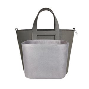 Puede incluir: Un bolso tote gris con un organizador interior gris a juego. El bolso tiene un diseño estructurado con un asa superior y una correa para el hombro desmontable. El organizador interior es un inserto rectangular suave.