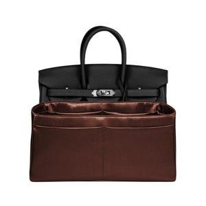 Könnte beinhalten: Eine schwarze Lederhandtasche mit silberfarbenem Verschluss und oberem Griff. Ein brauner Organizer mit mehreren Fächern befindet sich im Inneren. Die Handtasche ist ein klassischer Stil.
