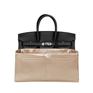 Può includere: Una borsa in pelle nera con due manici superiori e una chiusura in metallo color argento, posizionata dietro un organizer per borsa rettangolare in tessuto oro metallizzato con molteplici scomparti interni e design a giorno.