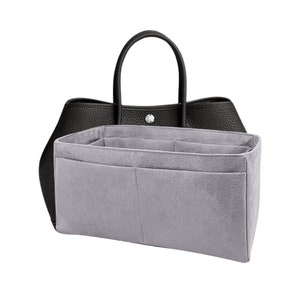 Peut inclure: Sac à main en cuir noir avec une poignée supérieure et un fermoir argenté. Un organisateur en feutre gris avec plusieurs compartiments est à l'intérieur, conçu pour ranger les objets. Le sac a une forme structurée et une texture grainée.