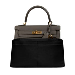 May include: A gray leather handbag with a top handle and gold-colored hardware. A black fabric organizer is placed inside the bag. The bag has a flap closure and a small leather tag attached to the handle.