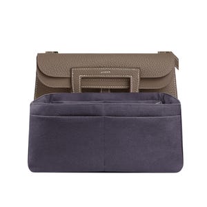 May include: A taupe leather handbag with a structured design, featuring a top handle and a flap closure. The bag has white stitching details and a dark blue interior organizer with multiple compartments. The handbag is designed to hold items.