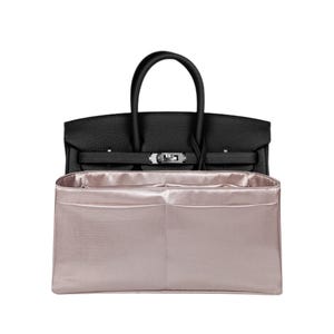 May include: A black leather handbag with a silver-toned metal clasp and two top handles, positioned behind a rectangular, metallic light pink fabric bag organiser insert featuring multiple interior compartments and a structured, open-top design.
