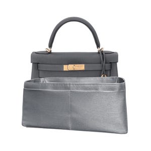May include: A grey leather handbag with a top handle and gold-coloured hardware. The bag has a structured design and a matching grey interior organiser. The bag is set against a white background.