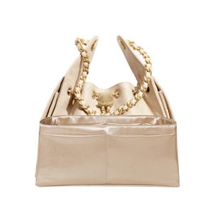 May include: Cream-colored handbag with gold chain straps and a decorative gold emblem. The interior features a matching cream-colored lining with multiple compartments. The bag is open, showcasing its spacious design and elegant details.