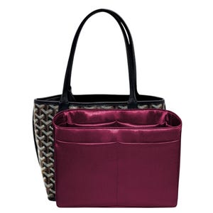 May include: A black and brown patterned tote bag with two thin black handles, paired with a rectangular, deep magenta satin bag organizer insert featuring multiple interior compartments and a smooth, reflective finish on the exterior surface.