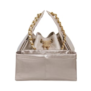 Puede incluir: Un bolso tipo cubo acolchado beige con correas de cadena doradas. El bolso tiene un cierre dorado y un organizador interior beige a juego con m&uacute;ltiples bolsillos. El bolso est&aacute; dise&ntilde;ado para llevarse por las correas de cadena.