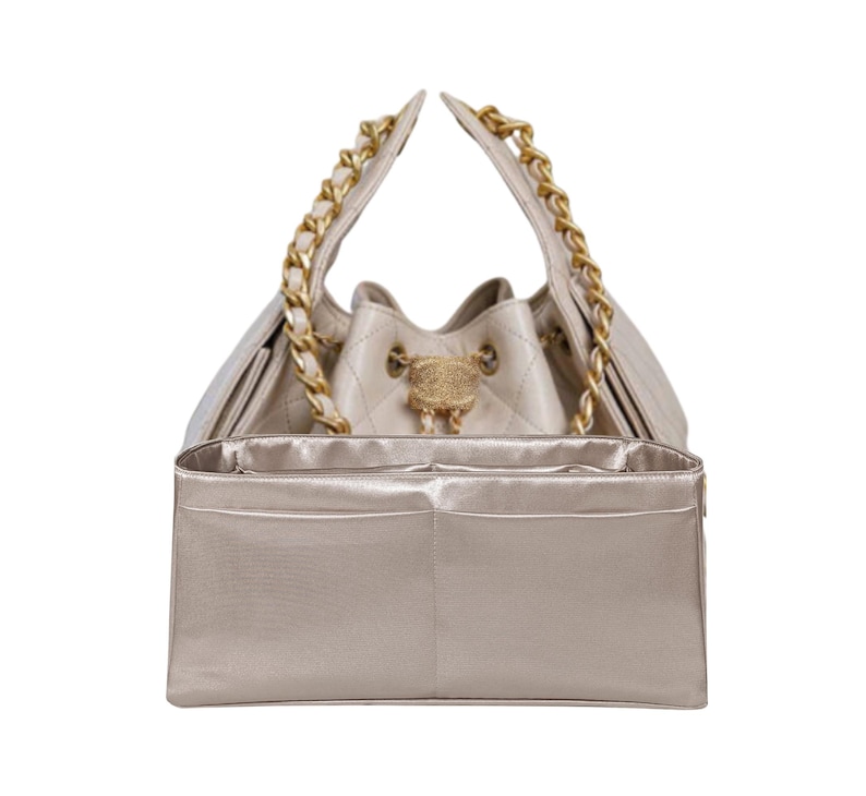 Puede incluir: Un bolso tipo cubo acolchado beige con correas de cadena doradas. El bolso tiene un cierre dorado y un organizador interior beige a juego con m&uacute;ltiples bolsillos. El bolso est&aacute; dise&ntilde;ado para llevarse por las correas de cadena.