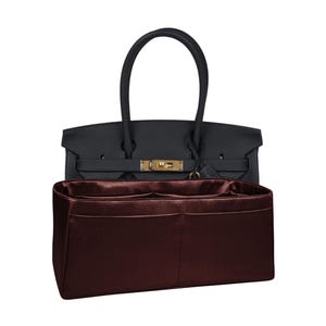 May include: A black leather handbag with a gold-tone clasp and handles. A burgundy-colored organizer is inside the bag, featuring multiple compartments. The bag is designed for carrying essentials and personal items.