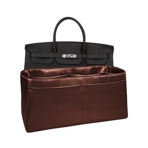 Könnte beinhalten: Eine schwarze Ledertasche mit silberfarbenen Beschlägen und ein rechteckiger Taschenorganizer aus metallisch bronzefarbenem Stoff mit mehreren Innenfächern und Taschen, der direkt vor der offenen Handtasche platziert ist.