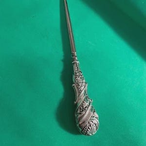 Peut inclure: Un bouton à crochet argenté antique avec une poignée ornée. La poignée présente des volutes détaillées et une forme bulbeuse, tandis que le crochet est fin et incurvé. Le bouton à crochet est posé sur un fond vert.