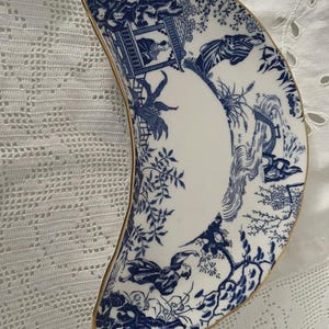 Könnte beinhalten: Eine halbmondförmige Porzellanschale mit blau-weißem Chinoiserie-Design. Die Schale hat einen goldenen Rand und zeigt eine detaillierte Landschaftsszene mit Figuren, Bäumen und architektonischen Elementen.