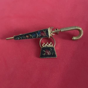 Könnte beinhalten: Ein Vintage-Broschen-Set in Goldoptik mit einem Regenschirm und einer Handtasche. Der Regenschirm hat ein schwarzes und floral gemustertes Verdeck und einen gewendelten Griff. Die Handtasche hat ebenfalls ein Blumenmuster. Das Set wird auf rotem Hintergrund präsentiert.