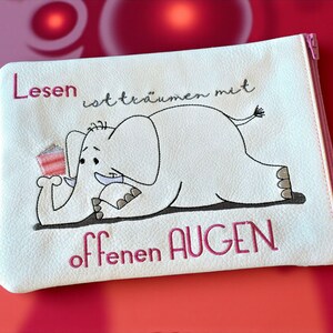 Könnte beinhalten: Eine weiße Reißverschlusstasche mit einer Cartoon-Elefanten-Illustration. Der Elefant hält ein Stück Kuchen. Der Text auf Deutsch lautet "Lesen ist träumen mit offenen AUGEN" in rosa Schrift. Der Reißverschluss ist rosa.