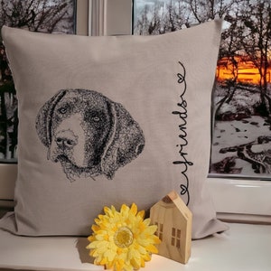 Könnte beinhalten: Beiges Zierkissen mit einer schwarzen Strichzeichnung eines Hundegesichts. Das Wort "friends" ist vertikal in Schreibschrift geschrieben. Eine gelbe Blume und ein kleines Holzhaus befinden sich vor dem Kissen.