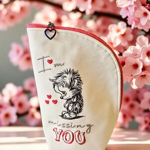 Könnte beinhalten: Eine weiße Kosmetiktasche mit rotem Reißverschluss und herzförmigem Anhänger. Die Tasche zeigt eine gestickte Illustration eines Igels und den Text "I'm missing YOU" in Rot und Grau.