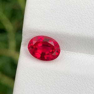 Könnte beinhalten: Ein facettierter, oval geschliffener Rubin. Der leuchtend rote Edelstein liegt auf einer weißen Oberfläche, mit einem verschwommenen grünen Hintergrund. Die Facetten des Edelsteins fangen das Licht ein und zeigen seine Brillanz und Klarheit. Eine Nahaufnahme zeigt die satte Farbe des Steins.