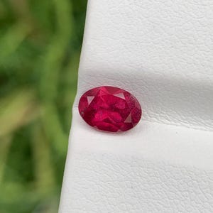 May include: A faceted, oval-shaped ruby gemstone. The ruby is a deep, vibrant red color and is set against a white background. The gemstone is approximately 0.5 inches in length.